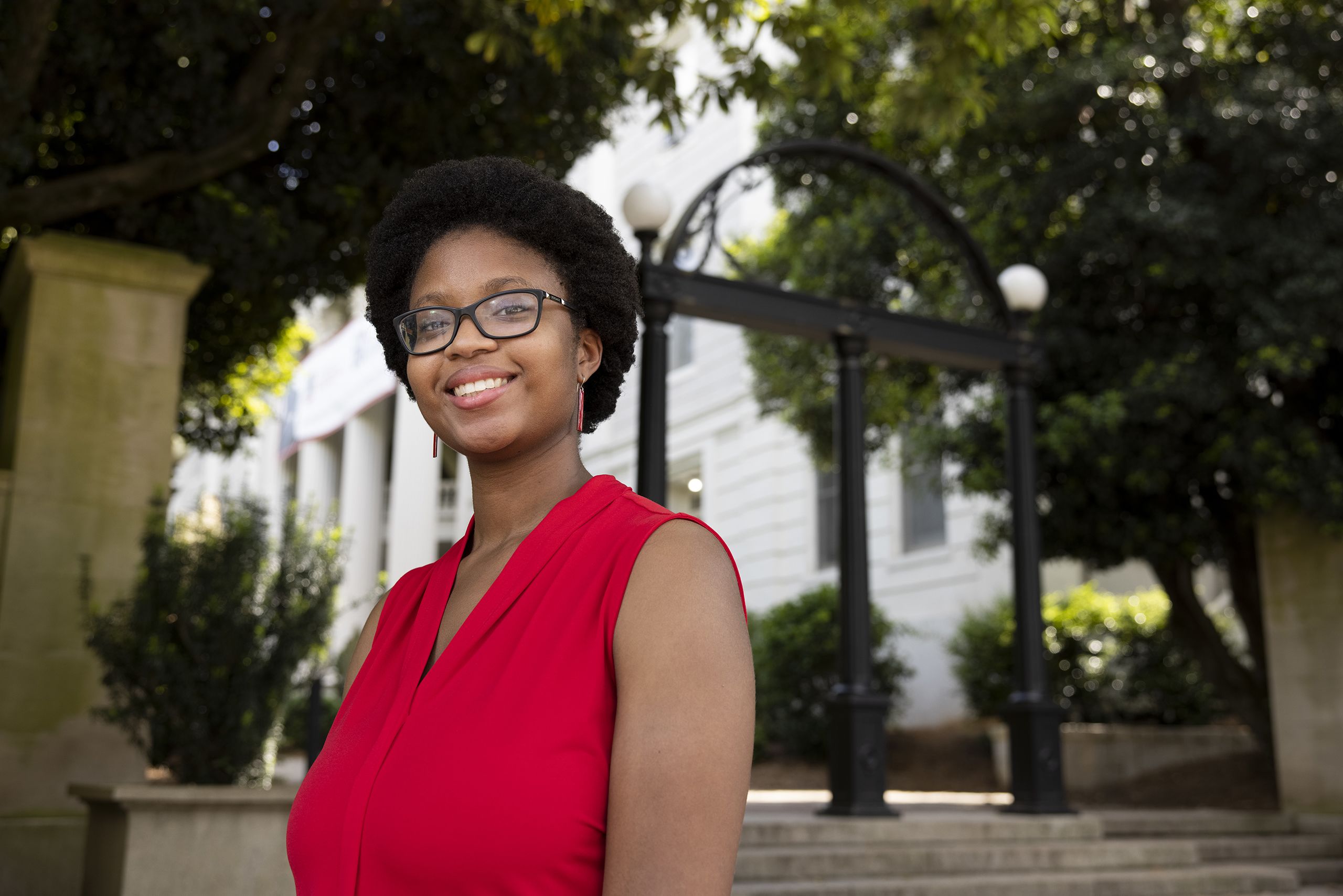 Phaidra Buchanan, wearing a red blouse, stands in front of the Arch on a sunny day in April 2021. 