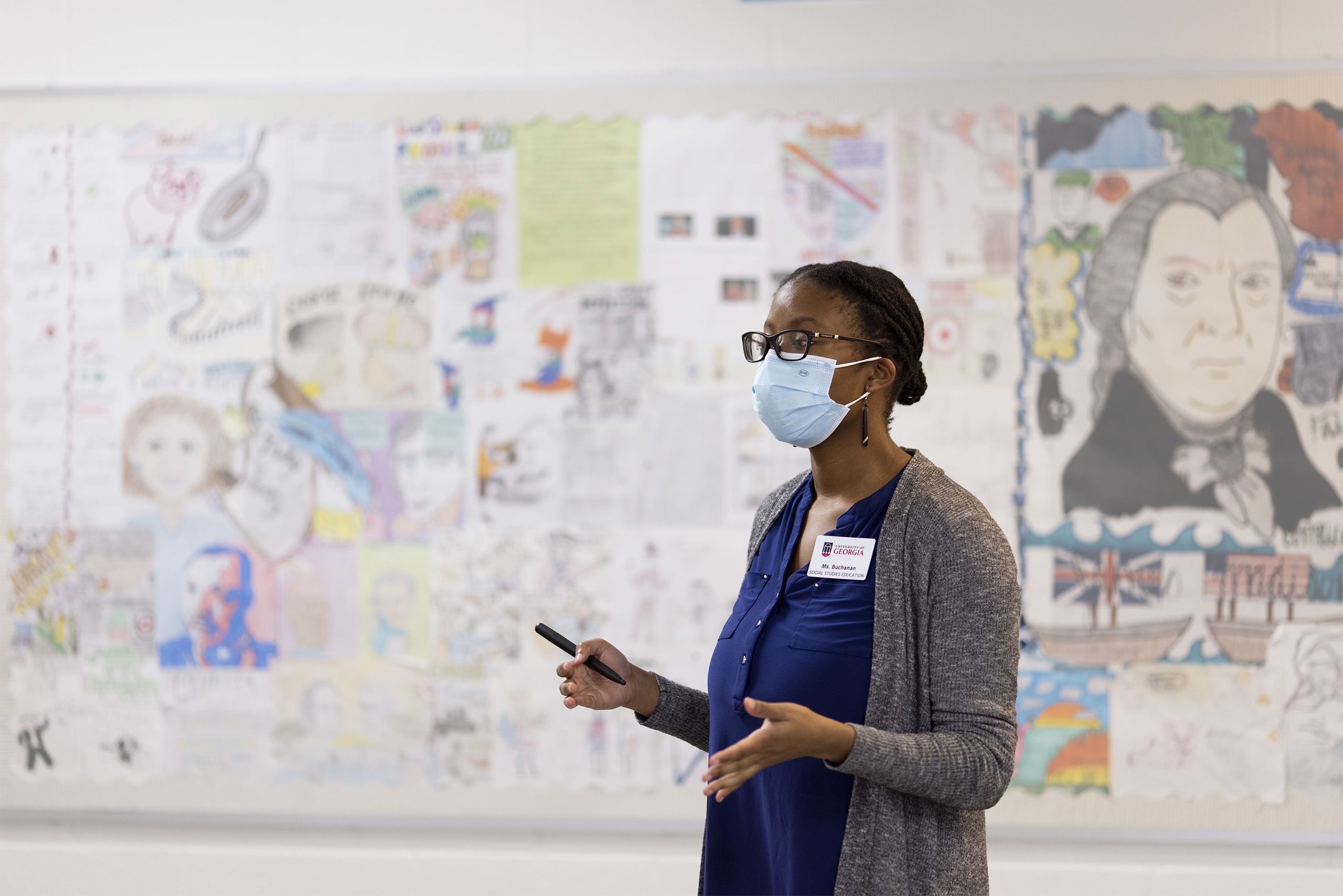 Phaidra Buchnanan stands in front of a colorful wall of student artwork depicting historical figures. She is wearing a face mask to prevent the spread of Covid-19 and lecturing. 