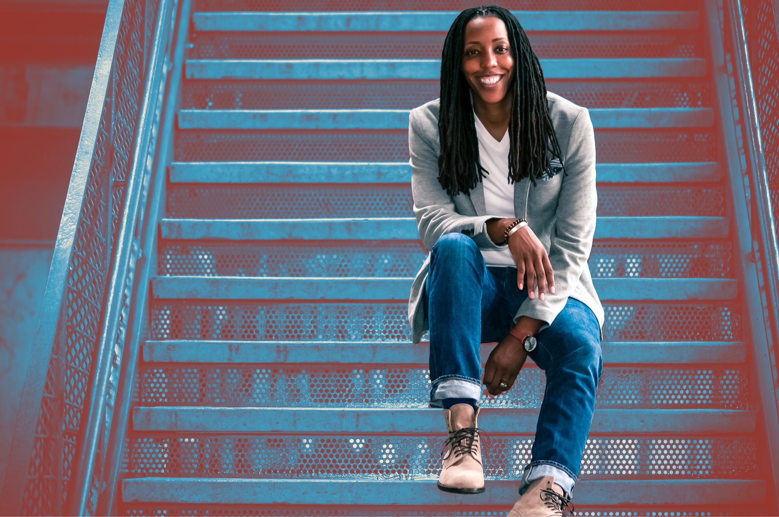 Professor Bettina Love, wearing jeans and a blazer, sits on a set of industrial metal steps and smiles at the camera. 